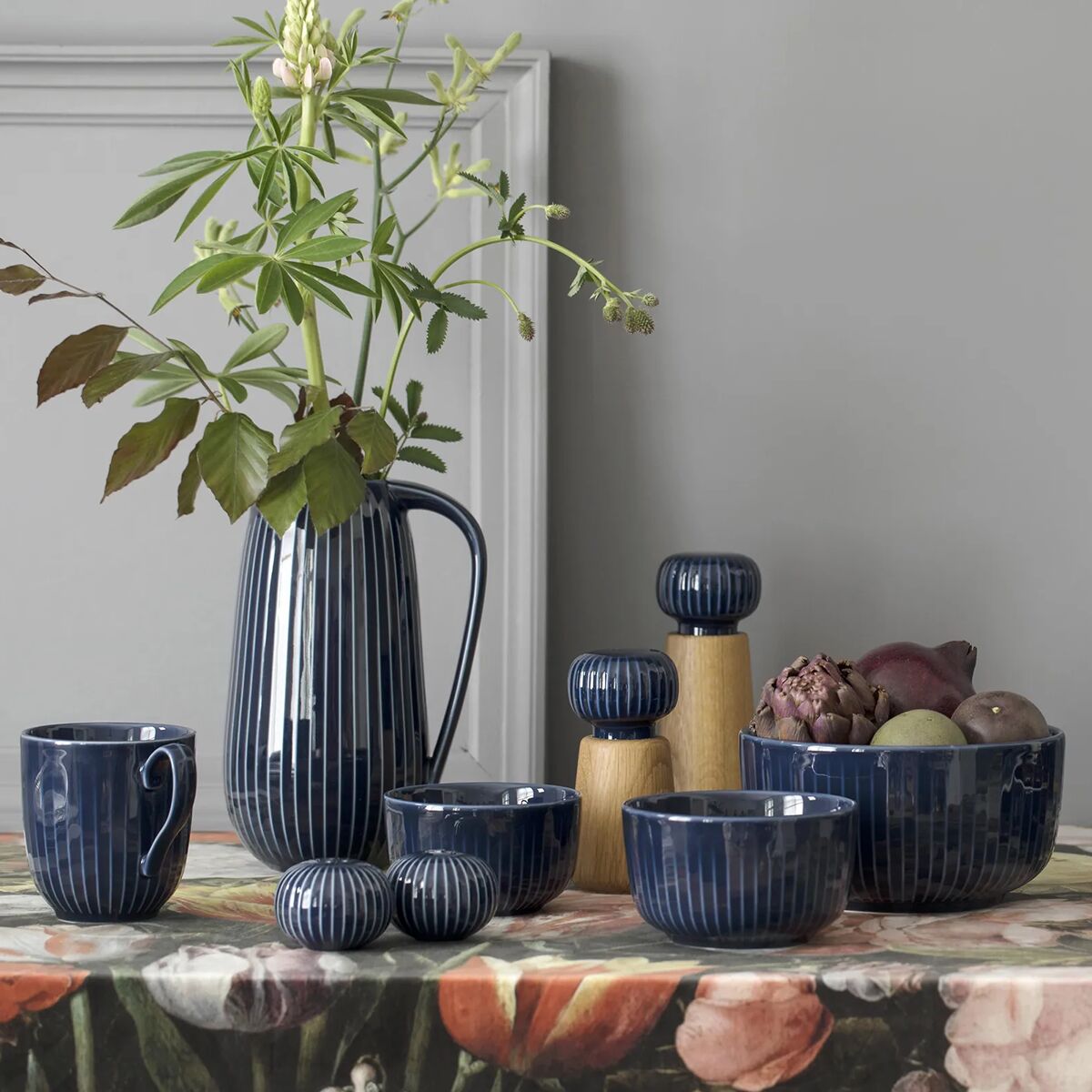 Die Abbildung zeigt einen gedeckten Tisch mit mehreren Keramikartikeln der Serie »Hammershøi«, darunter zwei Gewürzmühlen mit blauen Streifen und Holzelementen. Passend dazu sind Tassen, Schalen und eine Vase mit Blumen in denselben Blautönen angeordnet. Eine farbenfrohe Tischdecke mit Blumenmuster sowie frisches Obst und Gemüse runden das stilvolle Arrangement ab.