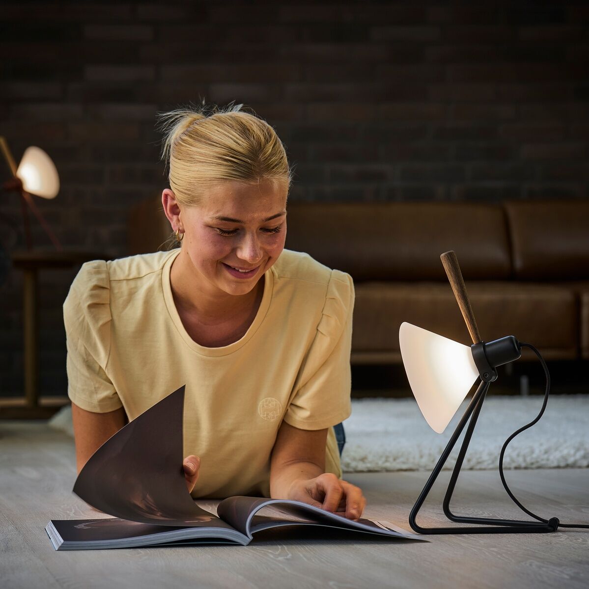 Eine Frau liegt auf dem Boden eines Wohnzimmers und liest ein Buch, das von der Tisch-/Hängelampe »Spot« beleuchtet wird. Die Lampe hat ein modernes Design mit einem schwarzen Metallrahmen und einem weißen Schirm. Im Hintergrund ist eine weitere Lampe in ähnlichem Design zu sehen, zusammen mit einer Couch und einem dunklen Ziegelwand.