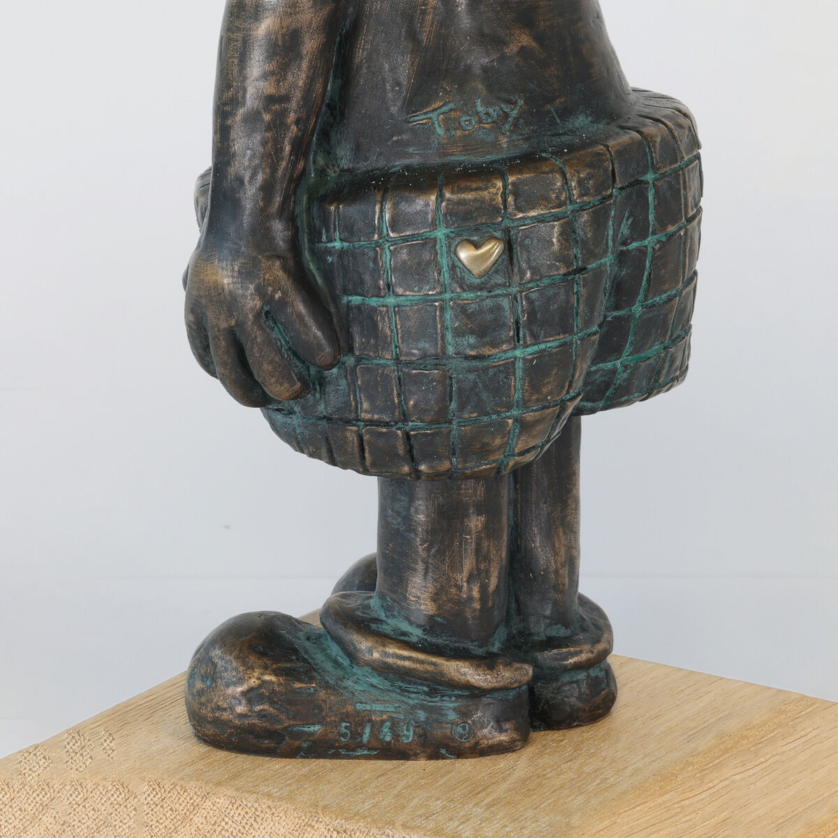 Detailansicht der Bronze-Holz-Skulptur »Guck maaal… für Dich!« von Toby Wichmann, Ars mundi, auf Holzsockel. Die Figur trägt karierte Shorts mit goldenem Herz, der Name des Künstlers und Nummerierung sind sichtbar. Oberfläche teils poliert, teils patiniert.