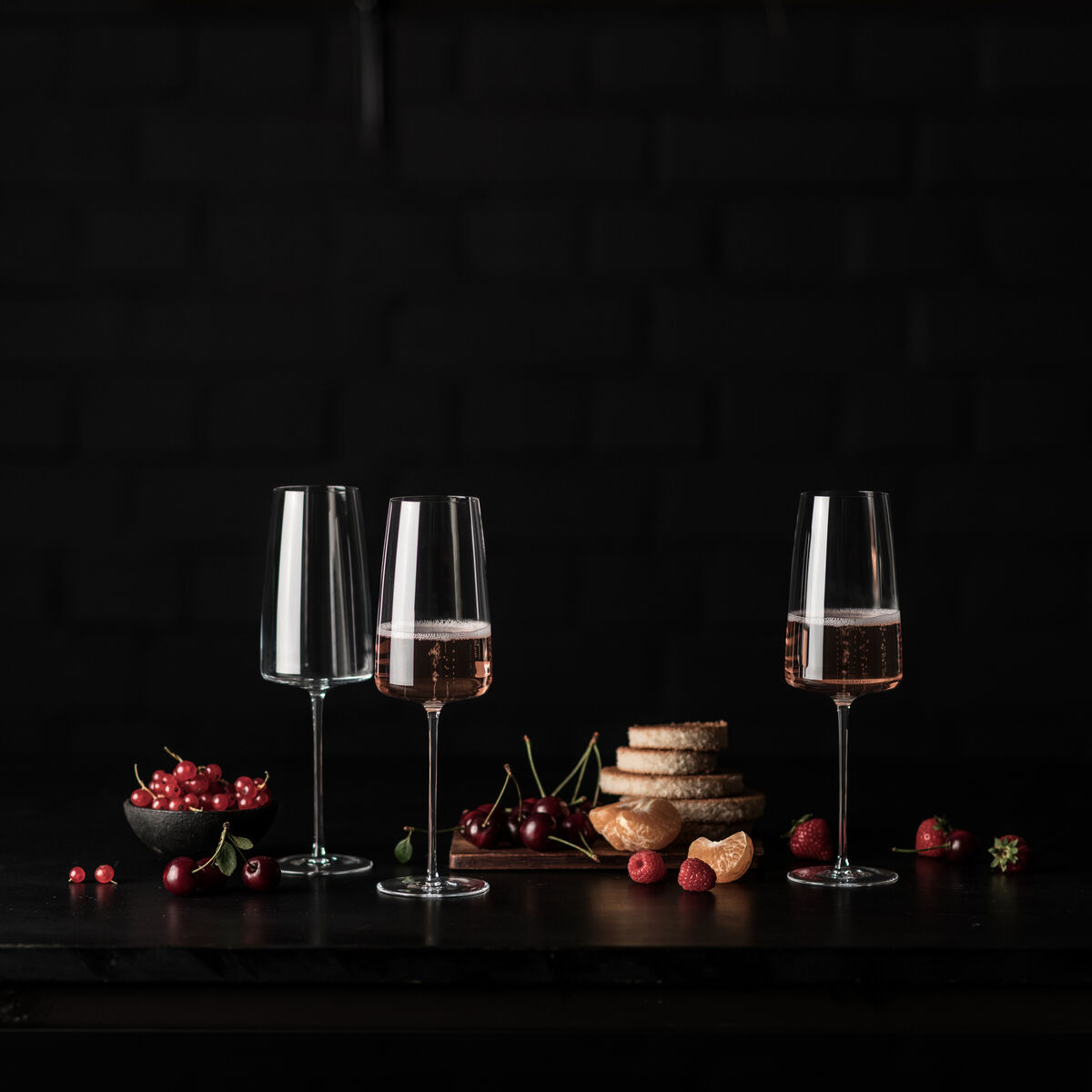 Zwei schlichte Sektgläser gefüllt mit roséfarbenem Getränk auf einem dunklen Tisch. Begleitet von ein paar frischen Kirschen und knusprigem Brot im Hintergrund. Dunkler Hintergrund, der die Gläser und die Früchte hervorhebt.