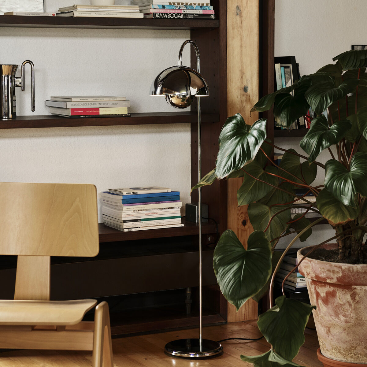 Ein stilvolles Wohnzimmer mit einer modernen Bodenlampe, einem Holztisch und einem großen Holzregal mit Büchern und Dekorationsgegenständen. Eine große Zimmerpflanze steht in einem Blumentopf neben dem Holztisch.