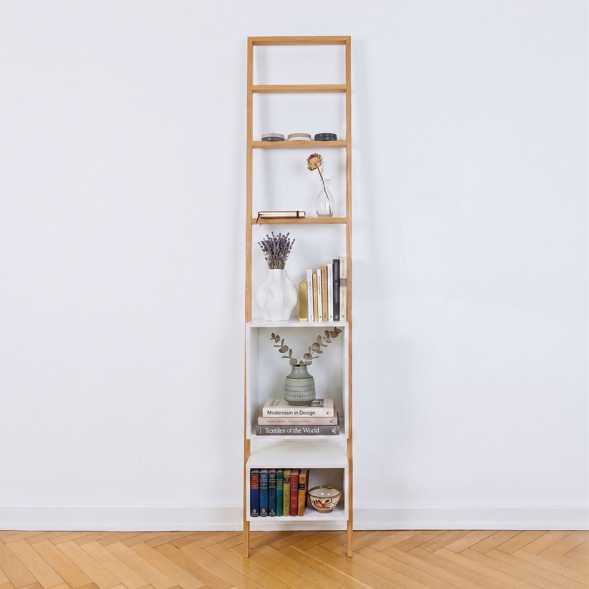 Schmales, an eine weiße Wand gelehntes Leiterregal aus geölter Eiche mit weißen Regalkästen. Es ist dekoriert mit Büchern, Vasen und Deko-Objekten. Der Boden ist aus hellem Echtholzparkett. Das Regal steht fest und wirkt modern sowie platzsparend.