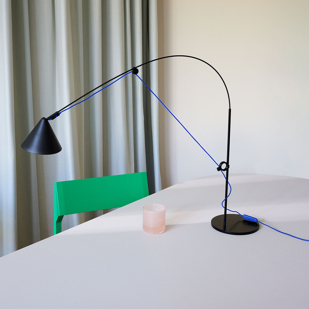 Eine moderne Tischlampe der Sonderedition AYNO mit einem minimalistischen Design. Die Lampe ist schwarz, mit einem flexiblen blauen Kabel und steht auf einem weißen Tisch. Im Hintergrund hängt ein beiger Vorhang. Eine grüne Stuhllehne ist ebenfalls sichtbar.