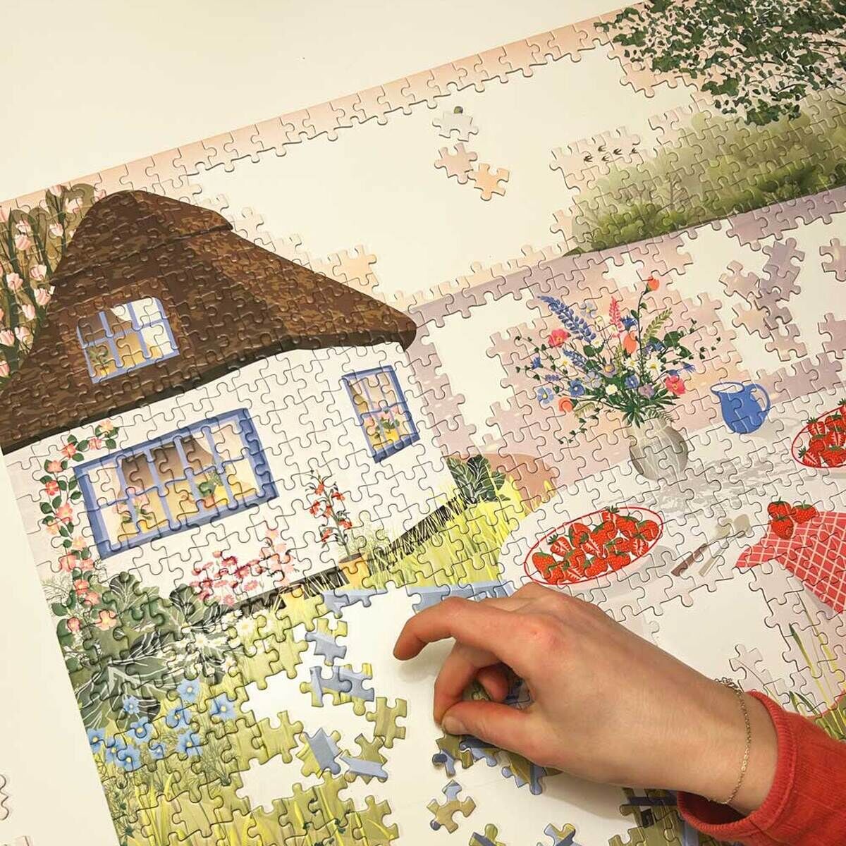 Ein fast fertig gestelltes Puzzle zeigt eine Szene mit einem weißen Haus mit braunem Dach im Hintergrund, einem Tisch mit roten und blauen Elementen im Vordergrund sowie Erdbeeren und Blumen in einem Garten. Eine Hand fügt gerade ein Puzzleteil ein.
