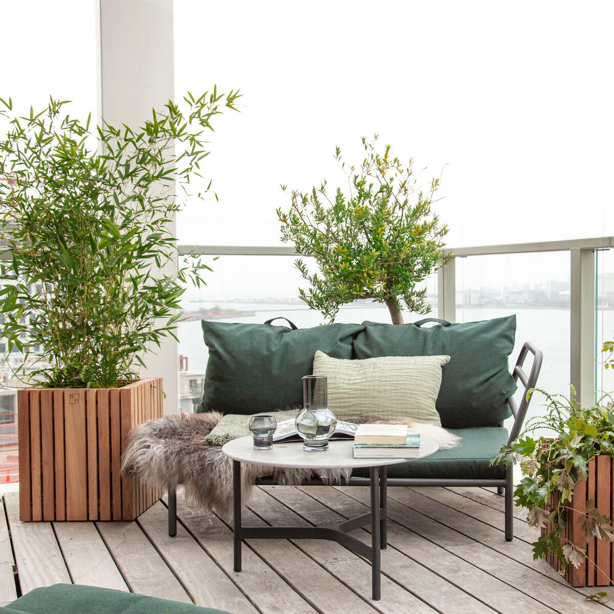 Eine gemütliche Terrasse mit modernen Pflanzgefäßen »GrowBIG«, die in Holzoptik gehalten sind. Es gibt mehrere grüne Pflanzen, die auf dem Boden und an einer Seite der Terrasse platziert sind. Ein kleiner rechteckiger Tisch mit einem Sofa und Kissen steht in der Mitte, umgeben von Pflanzen. Im Hintergrund ist eine Glasbrüstung, die Aussicht auf eine Stadt bietet.