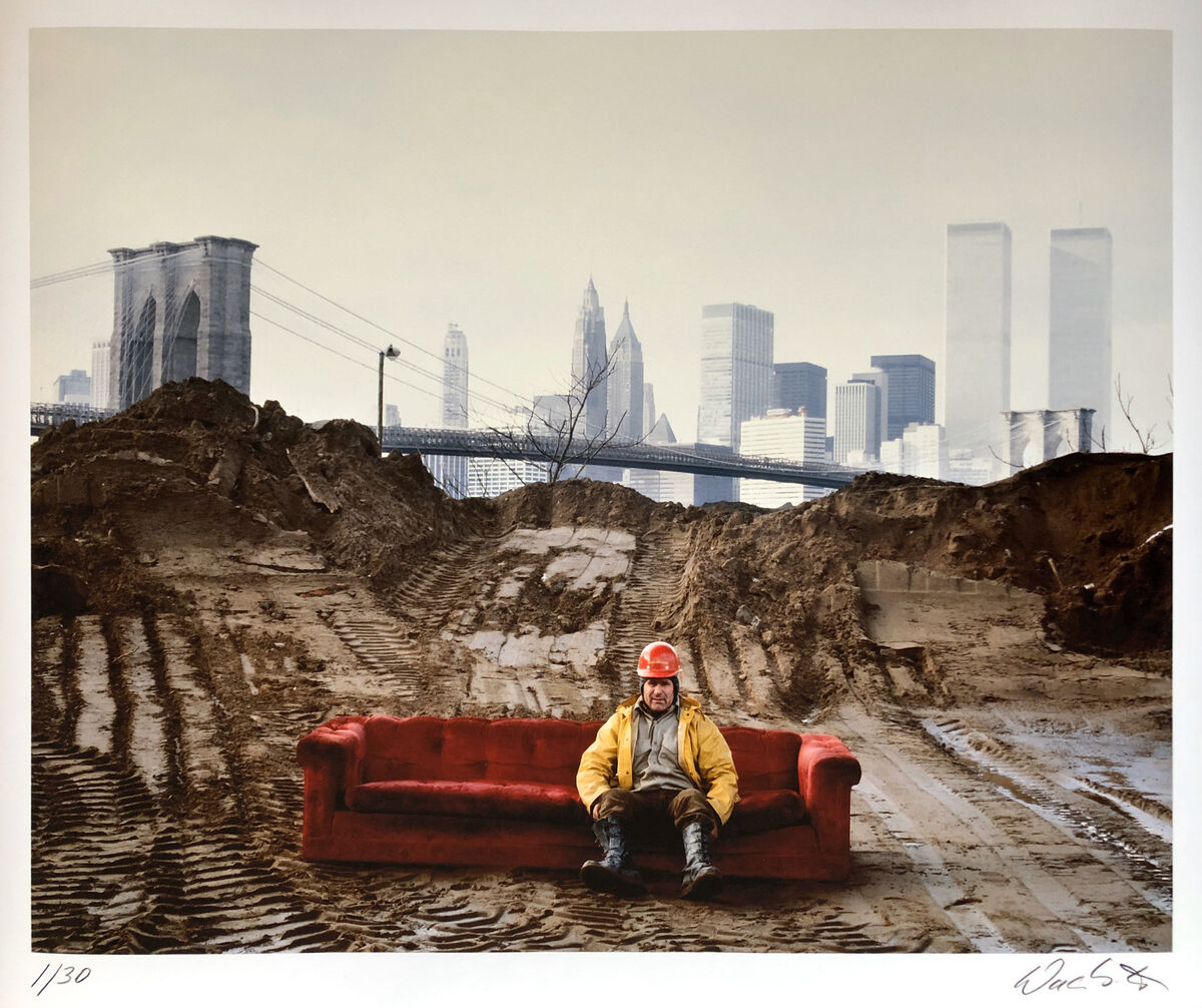 Farbfotografie von Wackerbarth aus dem Jahr 1979, betitelt »Peter Perniciaro«. Ein Mann sitzt auf einem roten Sofa auf einem mit Reifenspuren bedeckten Erdboden. Im Hintergrund sind städtische Elemente wie die Brooklyn Bridge und die Twin Towers zu sehen.