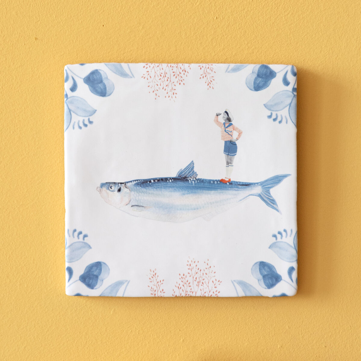 Keramikfliese mit einem Fischmotiv und einem kleinen menschlichen Charakter, auf einer gelben Wand montiert. Darunter sehen Sie zwei Keramikteller, einen blauen und einen orangefarbenen. Die Fliese hat einen weißen Hintergrund mit blauen und orangefarbenen Akzenten.