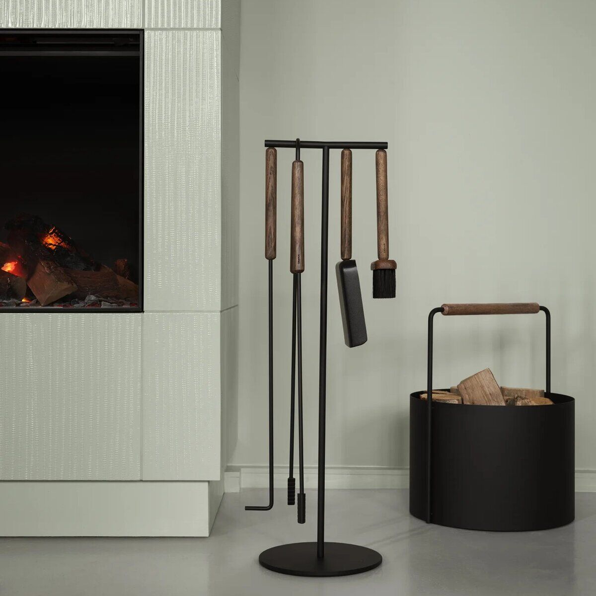 Modernes Kaminbesteck-Set 'ASHI' mit Griffen aus Holz und schwarzen Metallteilen steht neben einem Kamin mit loderndem Feuer, daneben ein schwarzer Holzkorb mit Holzscheiten, minimalistisches und skandinavisches Wohnambiente.