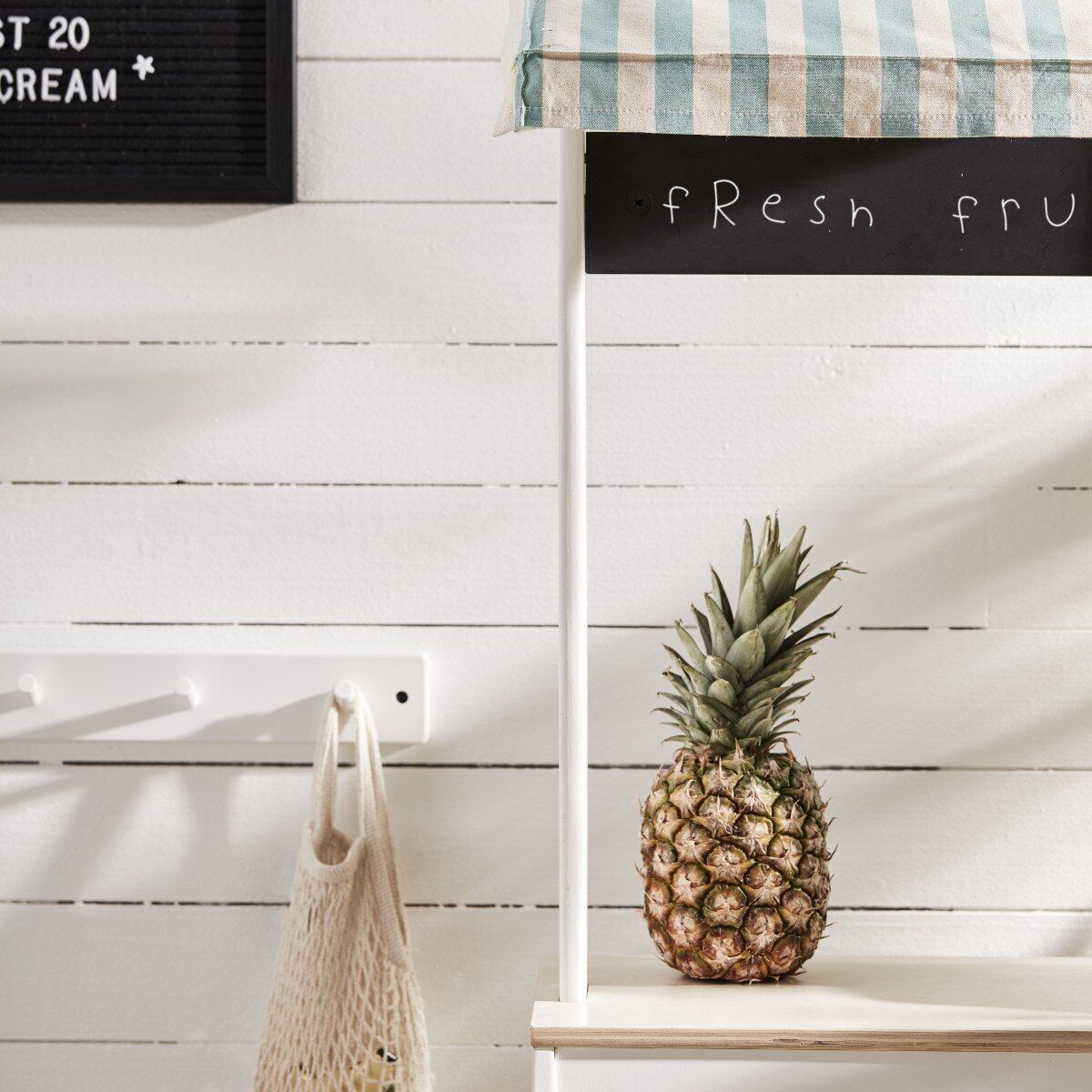Ein Marktstand für Kinder aus Holz mit einem blau-weiß gestreiften Stoffdach. Es gibt eine Anzeigetafel mit der Schrift 'Fresh Fruit' und einen einzelnen Ananas auf dem Stand.