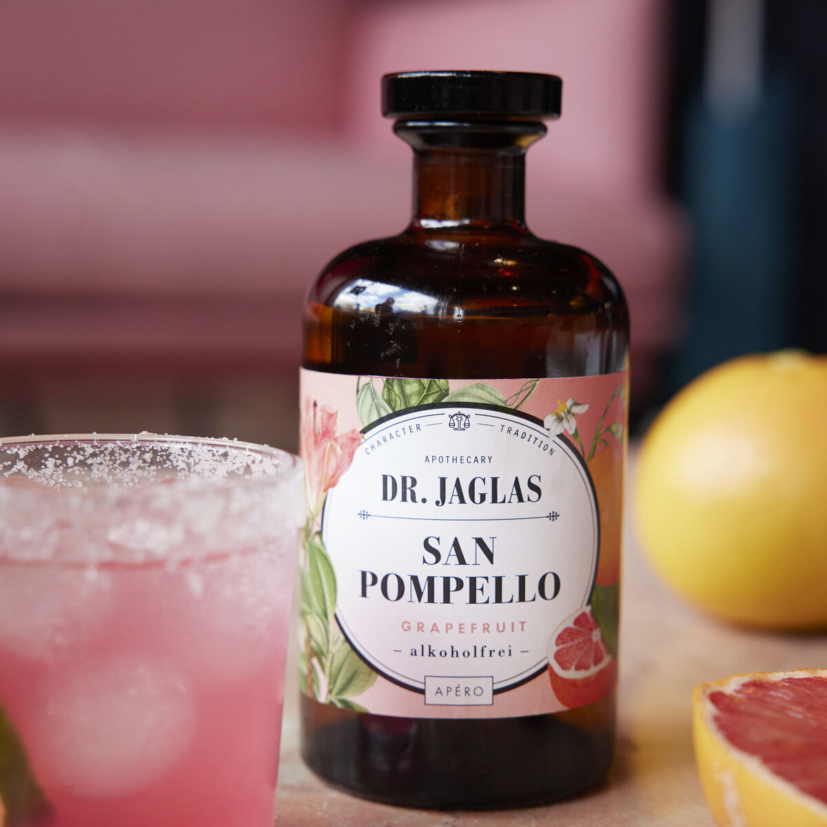 Eine braune Flasche »San Pomello« alkoholfreier Aperitif von Dr. Jaglas mit Grapefruit-Geschmack steht auf einem Tisch. Daneben ist ein Glas mit einem rosa Getränk und eine aufgeschnittene Grapefruit.