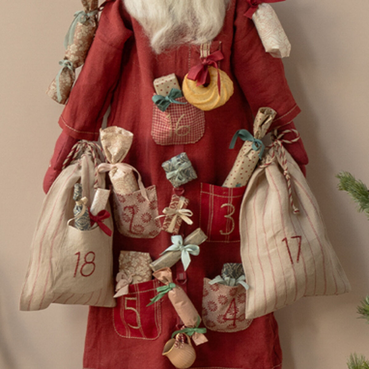 Ein adventskalender in Form eines freundlichen Weihnachtsmanns aus Stoff hängt an einer beigefarbenen Wand. Der Santa trägt einen roten Mantel und eine Mütze, an ihm sind nummerierte Beutel und kleine Geschenke befestigt. Daneben steht eine dekorative Metallbank mit eingepackten Geschenken und ein kleiner Tannenzweig. Die Szene verbreitet gemütliche Weihnachtsstimmung.
