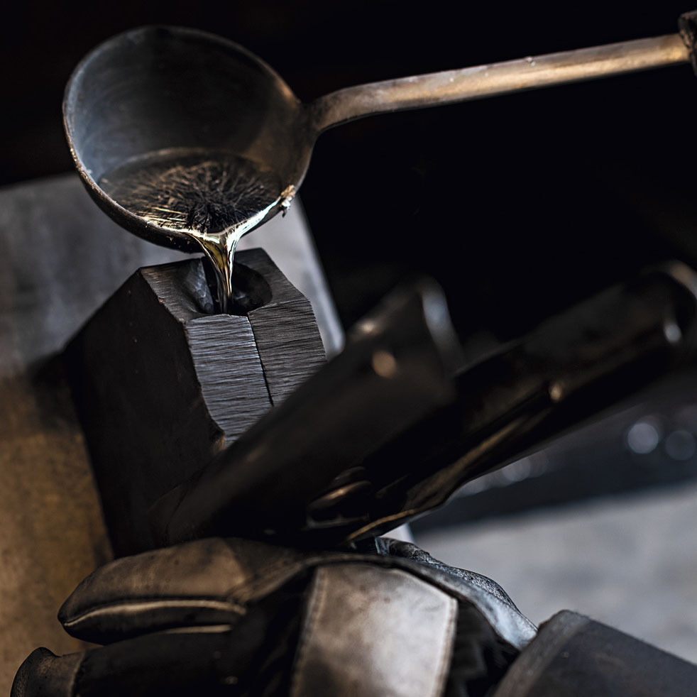 Detailaufnahme des Gussvorgangs: Schmelzflüssiges Zinn wird mit einer Kelle in eine Form gegossen. Im Vordergrund sind Hand und Schutzhandschuh des Handwerkers sichtbar. Die Szene wirkt traditionell und handwerklich geprägt.