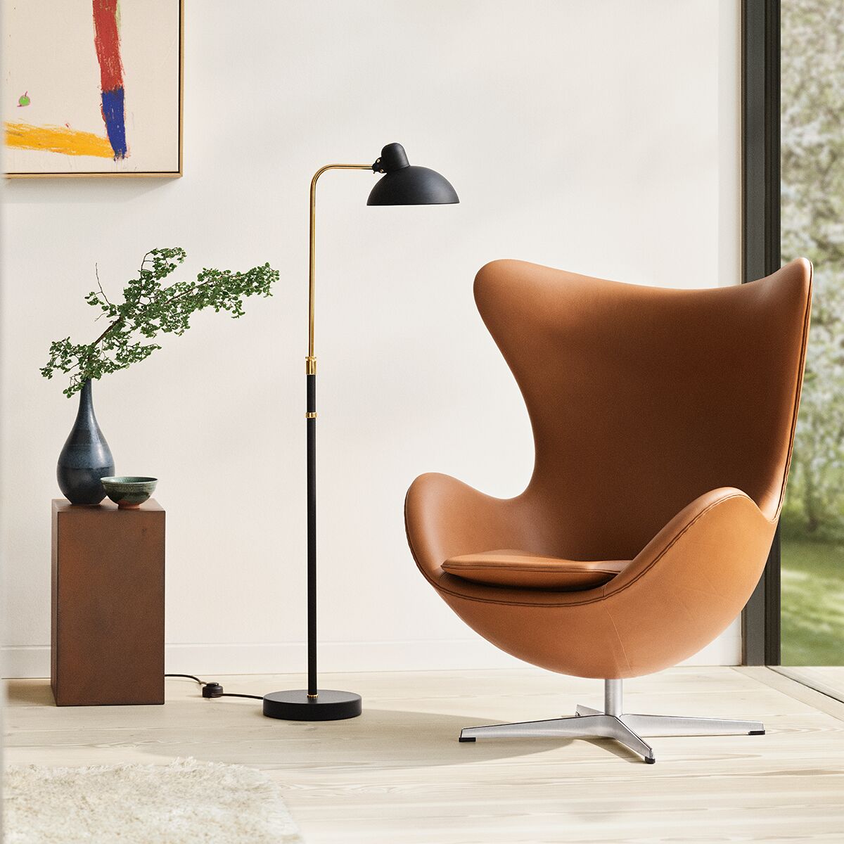 Modernes Wohnzimmer mit der Stehlampe »KAISER IDELL« matt von Fritz Hansen in Schwarz, schlank und elegant neben einem braunen Designersessel mit Metallfuß. Im Hintergrund eine kleine Vase mit grünem Zweig auf hohem Holzpodest, ein Fenster und ein abstraktes Bild an der Wand.