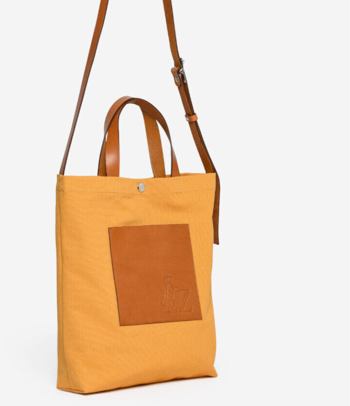 Eine modische Einkaufstasche aus dem ZEIT Shop mit dem Namen Jubiläums-Shopper. Die Tasche hat eine rechteckige Form, ist hauptsächlich gelb und hat braune Lederhenkel sowie einen verstellbaren langen Riemen. Auf der Vorderseite befindet sich eine kleine Tasche mit einer geprägten Zeichnung.