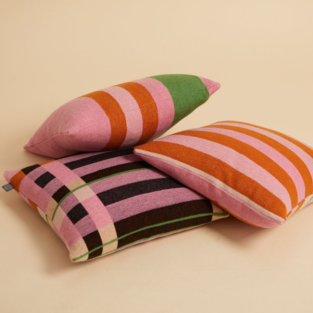 Drei Kissen mit Bauhaus-inspiriertem Design und geometrischen Mustern in den Farben Rosa, Orange, Grün, Schwarz und Weiß auf einem beige Hintergrund.