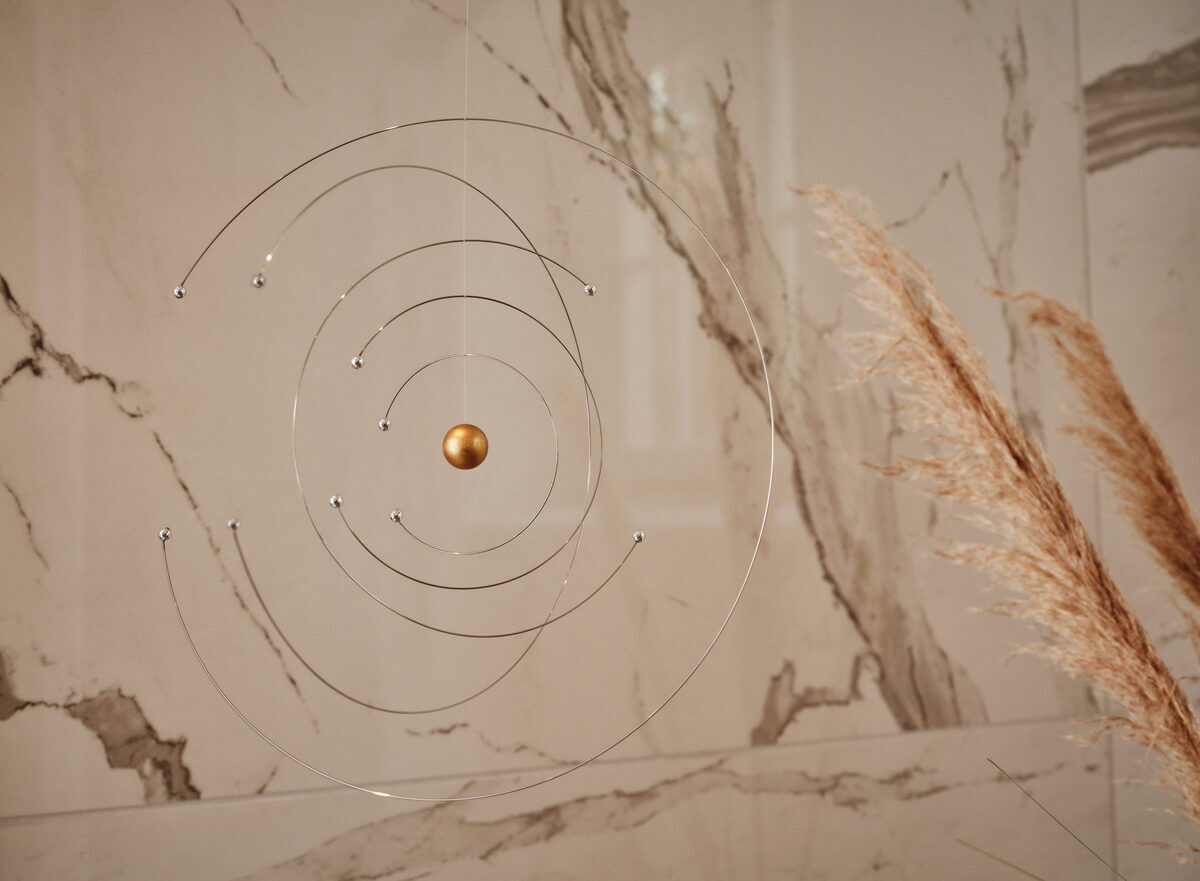 Ein minimalistisches Mobile, das Niels Bohrs Atommodell darstellt, hängt vor einem Marmor-Hintergrund. Dünne metallische Ringe und Kugeln in Gold und Silber bilden das Design. Daneben sind einige getrocknete Gräser zu sehen.