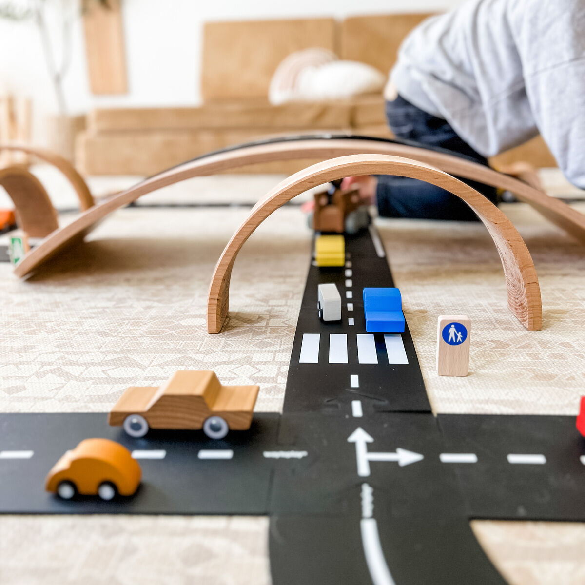 Ein Holzspielzeug-Straßenbau-Set mit aufgebauter schwarzer Straße, mehreren kleinen Holzautos, einem Fußgängerschild und bogenförmigen Holzbrücken auf einem hellen Teppich im Wohnzimmer. Ein Kind spielt im Hintergrund mit den Elementen.