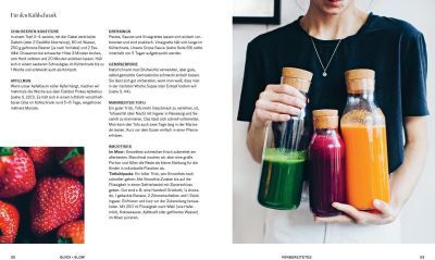 Eine Frau hält drei Glasflaschen mit gesunden grünen und roten Säften. Daneben steht ein aufgeschlagenes Kochbuch mit Rezepten und einem Foto von Erdbeeren.