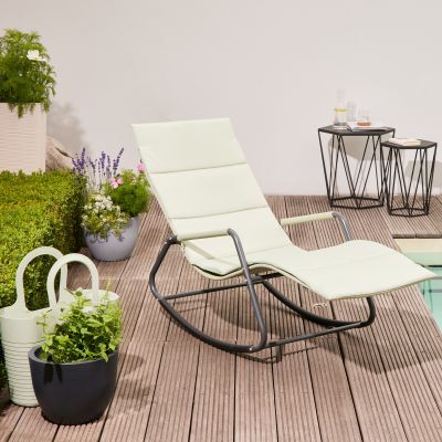 Eine moderne, cremeweiße Swingliege »Aliano« steht auf einer Holzterrasse neben einem Pool, umgeben von grünen Pflanzen und kleinen Beistelltischen mit Getränken. Badetuch und Gießkanne ergänzen die sommerliche Gartenszene.