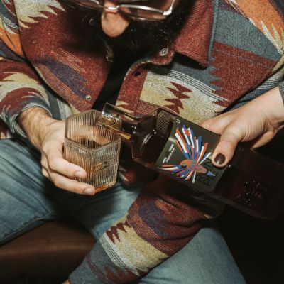 Eine Person in einer bunt gemusterten Jacke hält ein Glas, während eine andere Person eine Flasche Ron Piet Rum, 20 Jahre gereift, in das Glas einschenkt.
