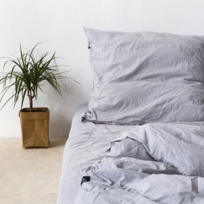 Eine gemütlich aussehende Bettwäsche in grauer Farbe auf einem Bett, daneben eine grüne Zimmerpflanze in einem braunen Topf. Die Wand im Hintergrund ist weiß.