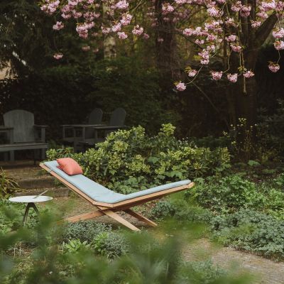 Eine stilvolle Holzliege steht in einem blühenden Garten mit einem blühenden Baum im Hintergrund. Die Liege ist modern gestaltet und liegt auf einem grünen Rasen. Es gibt einen kleinen weißen Tisch daneben. Die Szene strahlt eine ruhige und entspannende Atmosphäre aus.