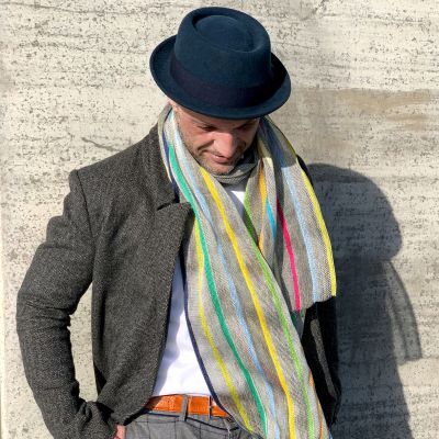 Ein länglicher Leinenschal mit naturweißer Grundfarbe liegt locker gefaltet. Quer verlaufen unregelmäßig bunte Streifen aus glänzender Seide in Rot, Grün, Blau, Gelb und Orange. Der Stoff wirkt leicht, luftig und hochwertig, mit feiner Struktur und dezentem Glanz.