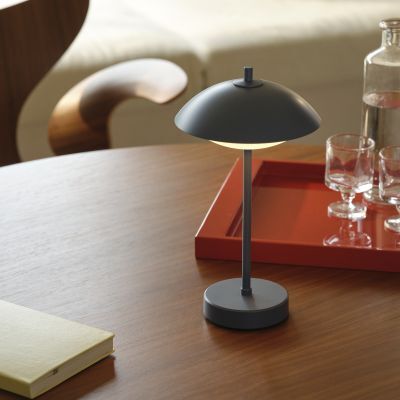 Auf einem runden Holztisch steht eine tragbare Lampe Clam™ von Fritz Hansen in mattem Blau. Daneben liegt ein gelbes Notizbuch. Im Hintergrund befindet sich ein rotes Tablett mit Gläsern und einer Glaskaraffe. Die Szene wirkt hell und stilvoll.